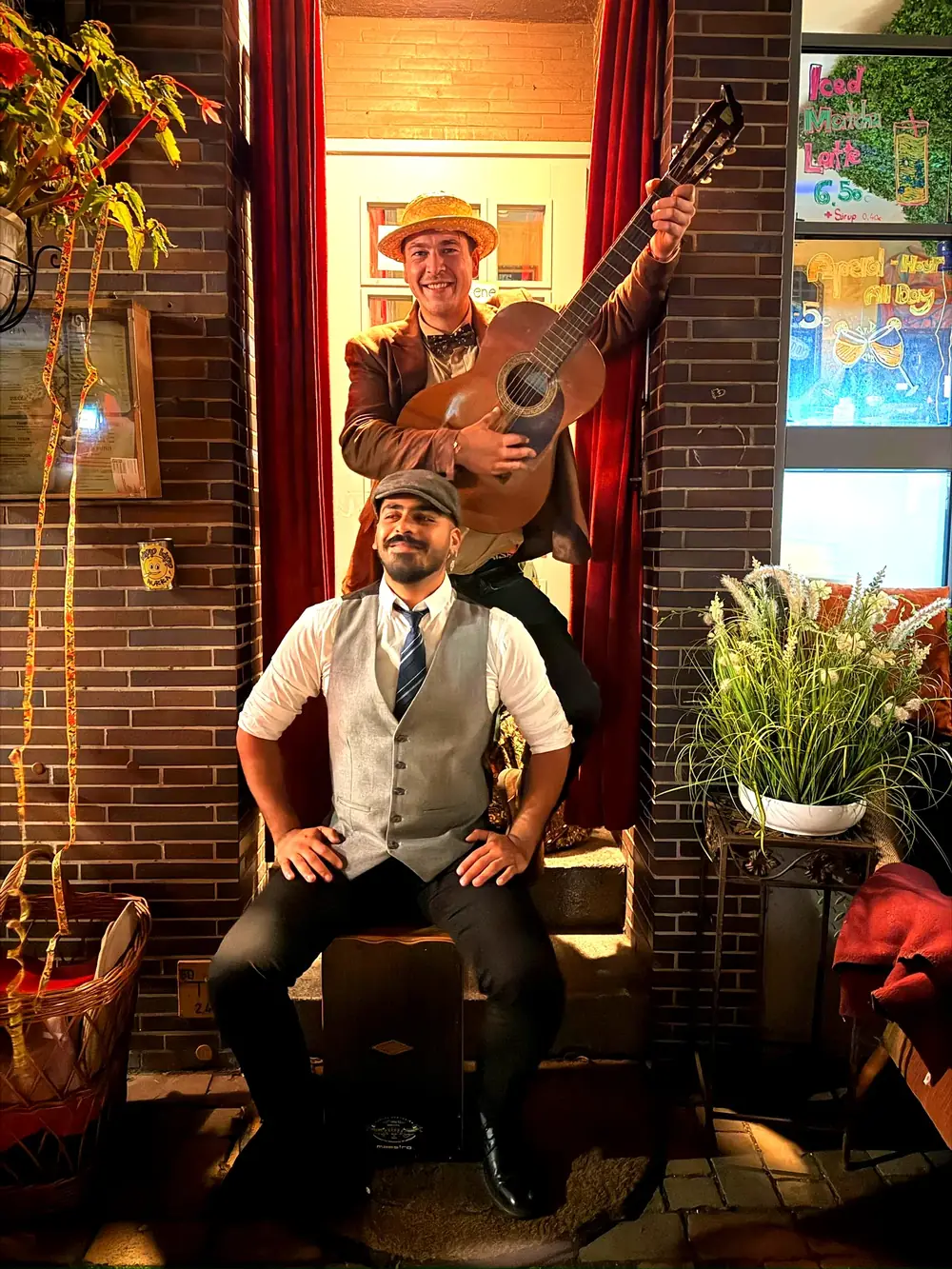 Ein Musiker mit Strohhut hebt die Gitarre, davor sitzt der andere mit Weste auf dem Cajón, vor einem Café bei Nacht